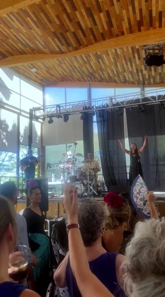 Palco interior com teto de madeira e grandes janelas, onde um trio atua com cantor de braços erguidos à direita, guitarrista à esquerda e baterista ao centro; o público em primeiro plano aplaude e levanta as mãos, num ambiente festivo que lembra um tríо com flamenquito e rumba para a tua boda em Córdoba.