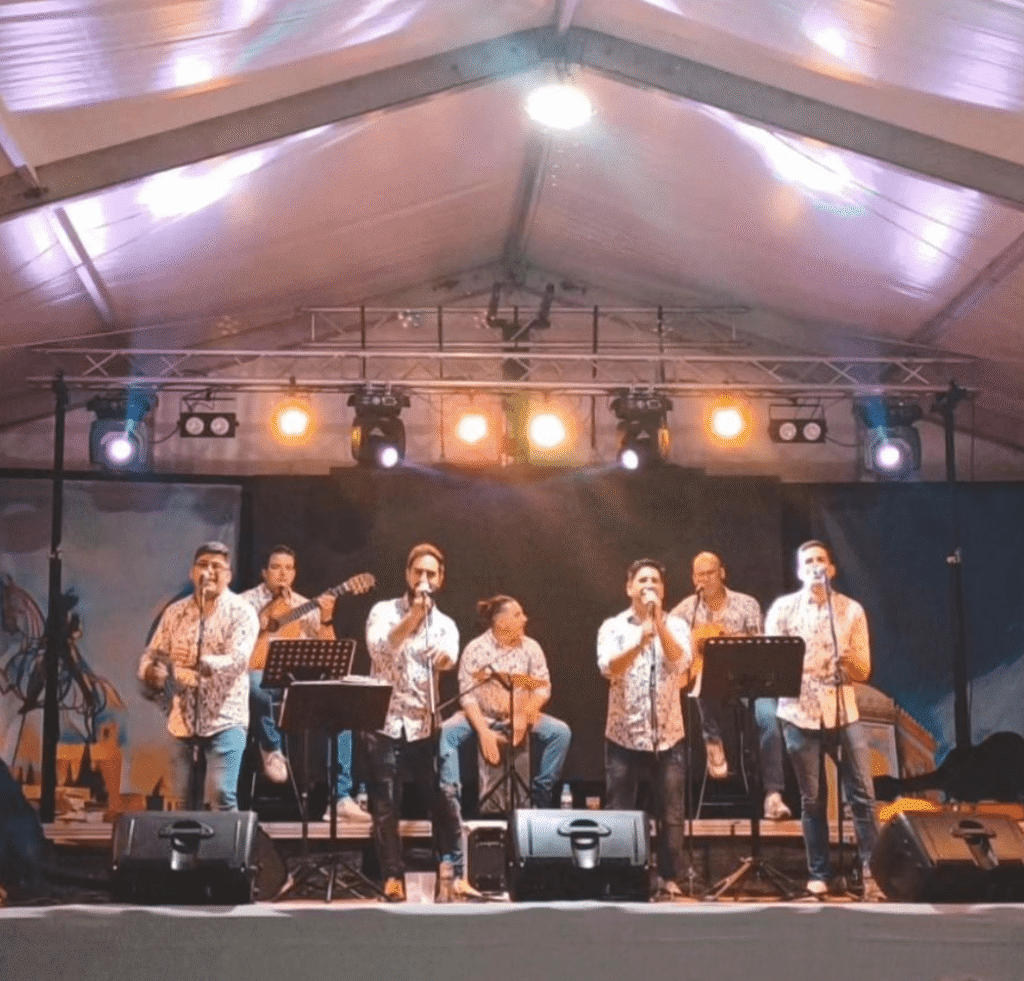 一个临时舞台上有七位音乐人面向观众表演，几位主唱手持麦克风，左侧有人弹木吉他，中间有人坐着打击乐器，舞台上方挂着多盏暖色灯。整体氛围热闹放松，适合用于巴达霍斯婚礼与私人庆典的西班牙派对音乐与现代弗拉门戈风格演出。