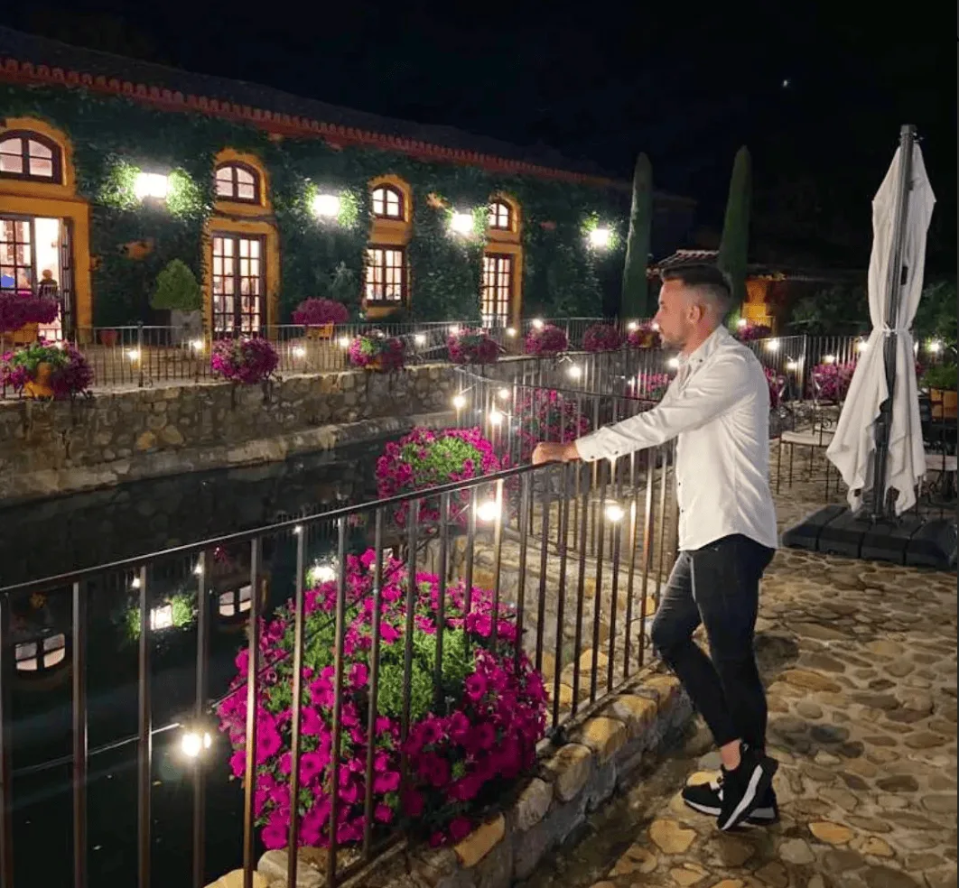 Hombre con camisa blanca apoyado en una barandilla, de noche, en un patio con luces cálidas, paredes cubiertas de hiedra y macetas de flores fucsias; una escena tranquila en un espacio de celebración, ideal para bodas y eventos en Barcelona con un dúo de flamenquito y rumba catalana.