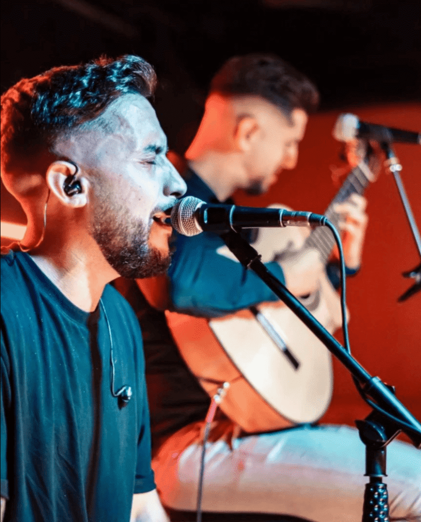 Cantor em primeiro plano a cantar num microfone com monitor intra auricular, enquanto ao fundo um guitarrista toca guitarra espanhola, num ambiente de palco com luz quente, ideal para um duo de flamenquito e rumba catalana para casamentos em Barcelona.