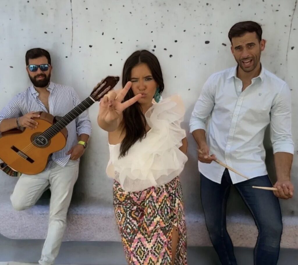 Tre musicisti in posa davanti a un muro chiaro macchiato: a sinistra un uomo con barba e occhiali da sole tiene una chitarra classica, al centro una donna con top bianco a balze fa il segno della vittoria verso la fotocamera, a destra un uomo in camicia bianca impugna due bacchette. Immagine promozionale di flamenquito e rumba fusion per matrimoni a Cordova in formato trio.