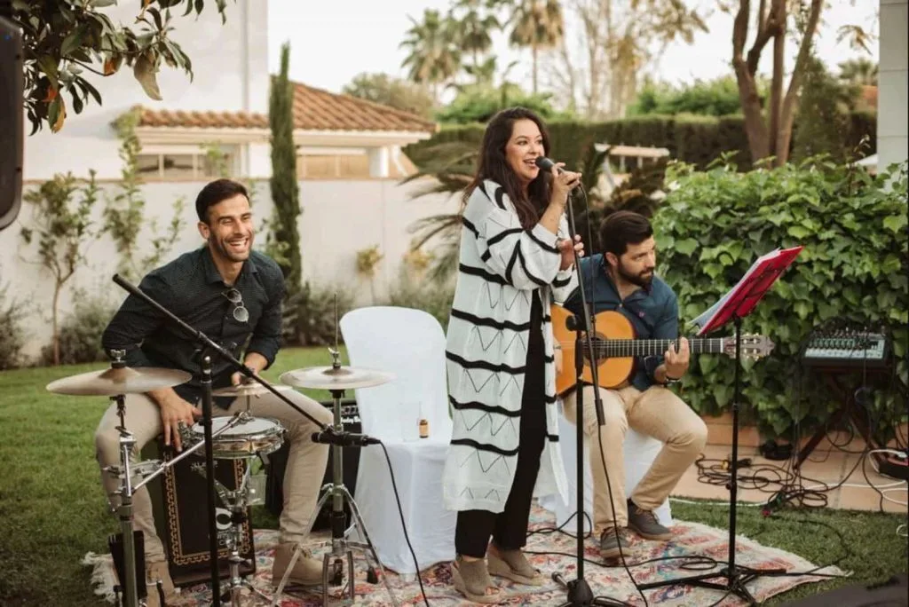 Trío musical en un jardín: cantante de pie con micrófono, guitarrista sentado tocando guitarra española y percusionista con cajón y platos; escena pensada para flamenquito y rumba fusión para bodas en Córdoba.