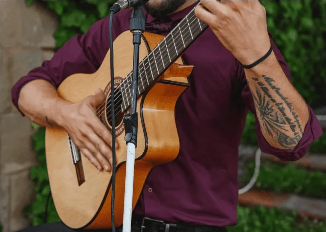 Primer plano de un músico tocando una guitarra española junto a un micrófono; se ven sus manos sobre las cuerdas y el mástil y parte del torso con camisa morada. Imagen asociada a un dúo de flamenquito y rumba catalana para bodas en Barcelona.