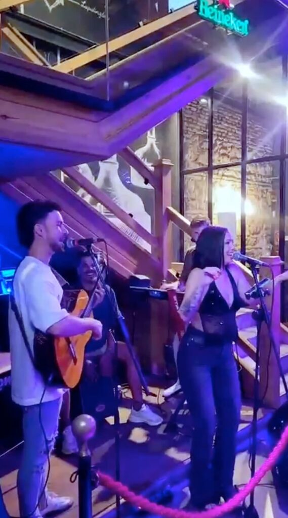 Binnen in een bar of eventlocatie staat een trio op een klein podium bij een houten trap: links een man die gitaar speelt en zingt in een microfoon, rechts een vrouw die zingt en met haar handen het publiek aanmoedigt, en achter hen een toetsenist. Het beeld past bij een pop flamenco trio in Córdoba dat sfeer brengt tijdens het namiddagfeest van een bruiloft, met paarse en blauwe verlichting en een Heineken bordje bovenin.