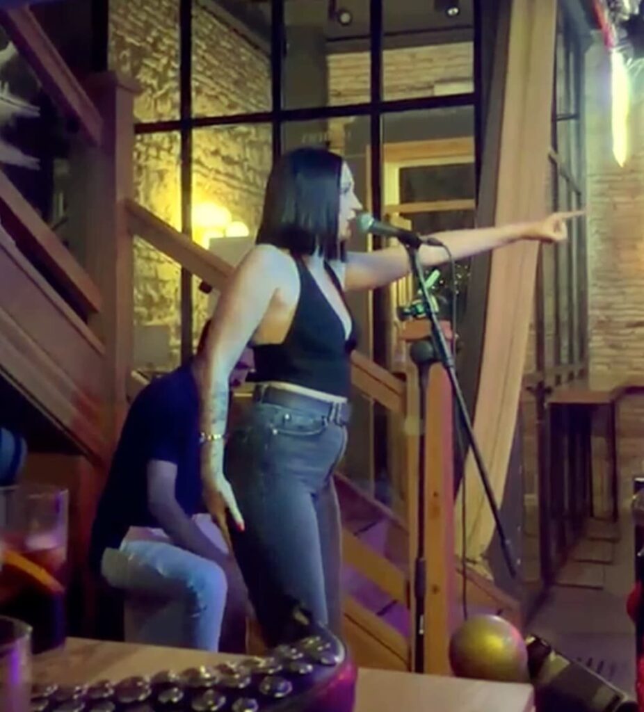 Zangeres staat zijwaarts bij een microfoon en wijst met gestrekte arm naar rechts, terwijl een muzikant achter haar zit in een warme binnenruimte met houten trap en grote ramen; sfeer van een popflamenco trio in Córdoba om het tardeo van een bruiloft extra leven te geven.