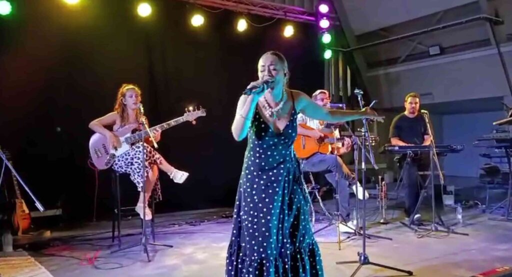 Palco com cantora ao centro, de vestido comprido com bolinhas, a cantar ao microfone enquanto uma baixista sentada toca à esquerda e um guitarrista toca atrás; luzes coloridas no topo e equipamento musical à volta, imagem usada para ilustrar um trio com flamenquito e rumba para a tua boda em Córdoba.