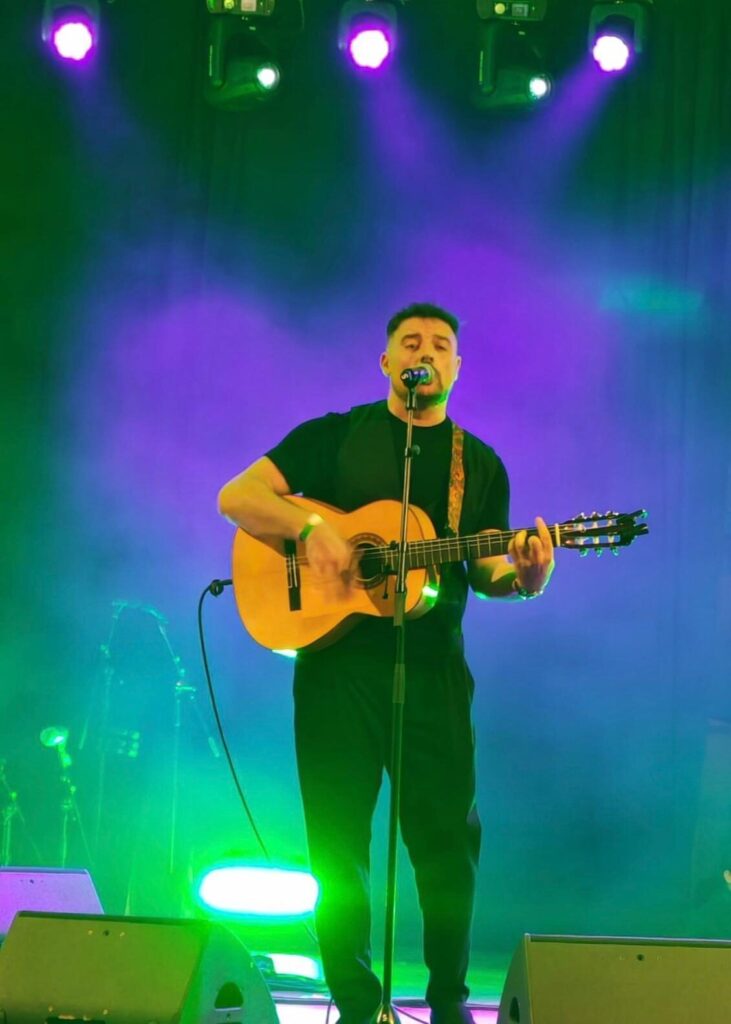 Cantant amb guitarra espanyola davant d’un micròfon, sota llums verdes i liles d’escenari; una imatge de rumba fusió a Barcelona per a casaments amb ganes de festa.
