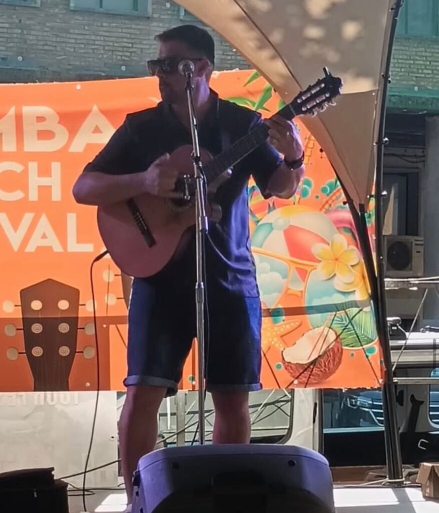 Un guitarrista amb ulleres de sol toca una guitarra espanyola davant d’un micròfon, en un escenari cobert amb una lona i un fons taronja decorat; una imatge de rumba fusió a Barcelona per a bodes amb ganes de festa.