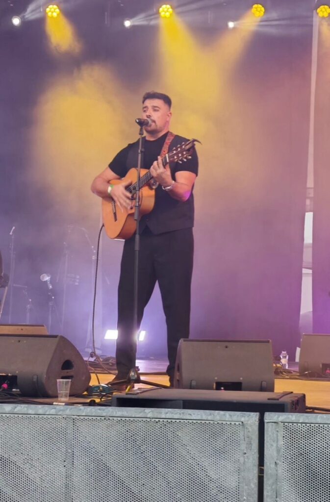 Músic sobre un escenari amb micròfon i guitarra acústica, cantant mentre toca sota focus grocs i una mica de fum, en un ambient de festa ideal per a rumba fusió a Barcelona per a casaments amb ganes de festa.