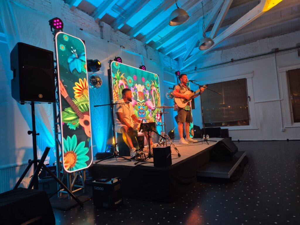 Escenari interior amb dos músics: un canta i toca la guitarra davant d’un micròfon, i l’altre està assegut amb percussió i equip de so al seu voltant. Al fons hi ha pantalles verticals amb dibuixos de flors i colors vius, i llums de focus que tenyeixen el sostre de blau, creant un ambient festiu ideal per a rumba fusió a Barcelona per a casaments amb ganes de festa.