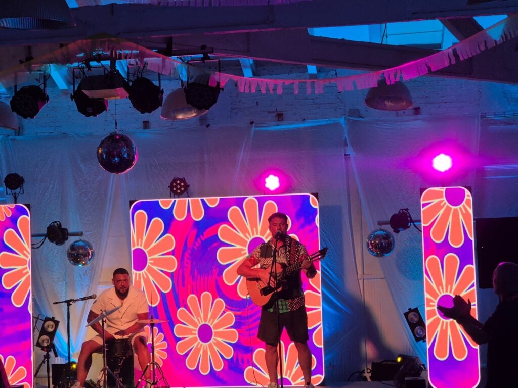 Escenari interior amb il·luminacio magenta i blava en una celebracio: un cantant toca la guitarra i canta al microfon al centre, i un percussionista seu a l esquerra amb un petit set de bateria. Al fons hi ha pantalles amb dibuixos de flors psicodeliques i boles de mirall penjades, creant un ambient festiu ideal per a rumba fusio a Barcelona per a casaments amb ganes de festa.