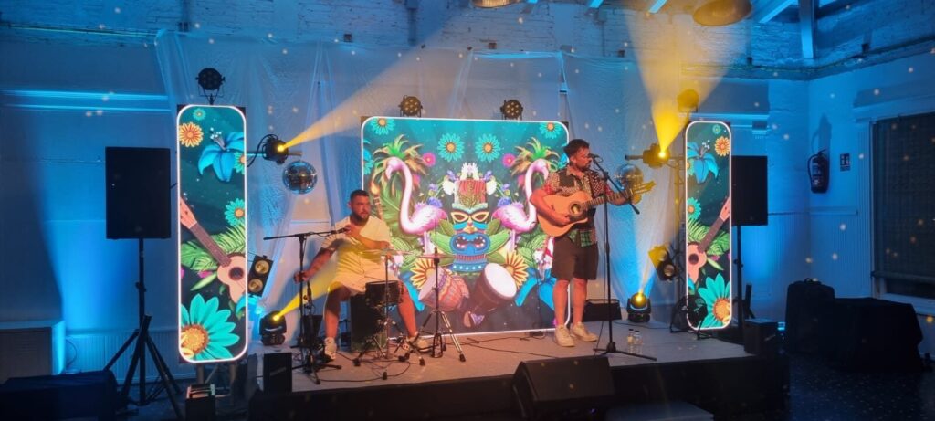 Dos músics sobre un escenari interior il·luminat amb llums blaves i grogues, un canta i toca la guitarra davant d’un micròfon i l’altre toca una bateria petita; al fons hi ha pantalles amb un disseny tropical amb flamencs i flors, ambient festiu ideal per a rumba fusió a Barcelona per a casaments amb ganes de festa.