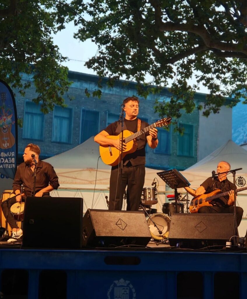 Tres músics sobre un escenari exterior, sota l’ombra d’arbres. Al centre hi ha un home dret tocant una guitarra acústica davant d’un micròfon. A l’esquerra, un percussionista assegut amb un petit tambor a les cames mira cap al costat. A la dreta, un baixista assegut toca un baix elèctric amb un atril de partitures al davant. Al fons es veuen carpes blanques i la façana d’un edifici. Imatge relacionada amb rumba fusió a Barcelona per a bodes amb ganes de festa.
