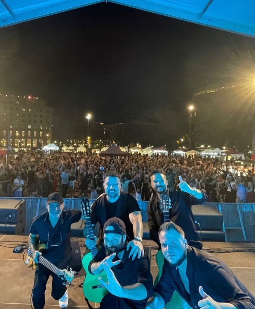 Cinc músics a sobre d’un escenari de nit posant per a una foto amb dues guitarres, mentre al fons es veu una plaça plena de públic i casetes iluminades; una imatge que transmet rumba fusió a Barcelona per a casaments amb ganes de festa.