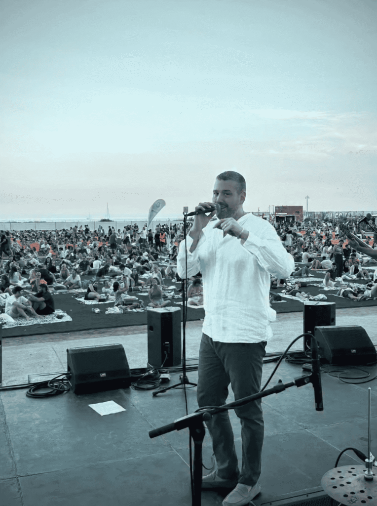Cantor em palco ao ar livre, de camisa branca, a segurar um microfone e a falar para um grande público sentado no chão, com colunas de som e suportes de microfone à frente; ambiente de festa popular na orla, ideal para um trio de pop espanhol e rumba para festas maiores em Barcelona.