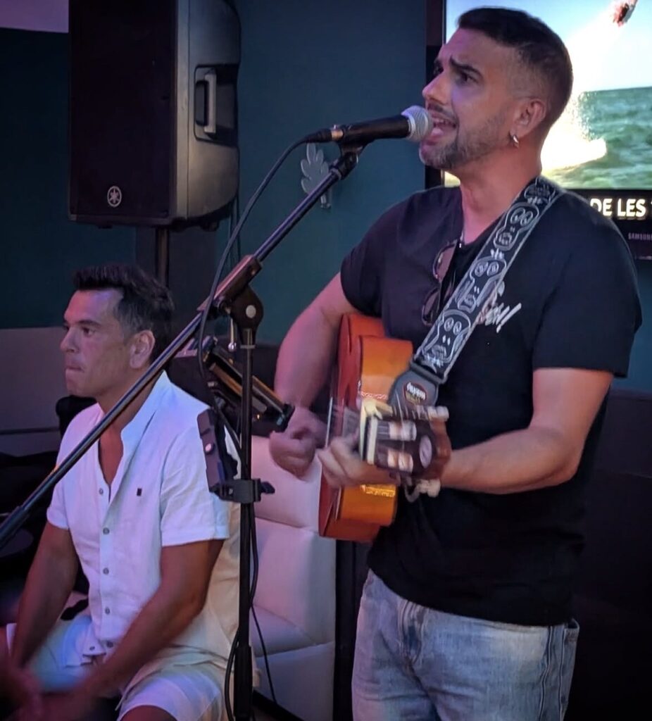 Cantor e guitarrista a tocar uma guitarra espanhola e a cantar num microfone, com outro músico sentado ao lado, num espaço interior com colunas de som; ambiente de rumba fusão e flamenquito para um aniversário de verão em Barcelona.