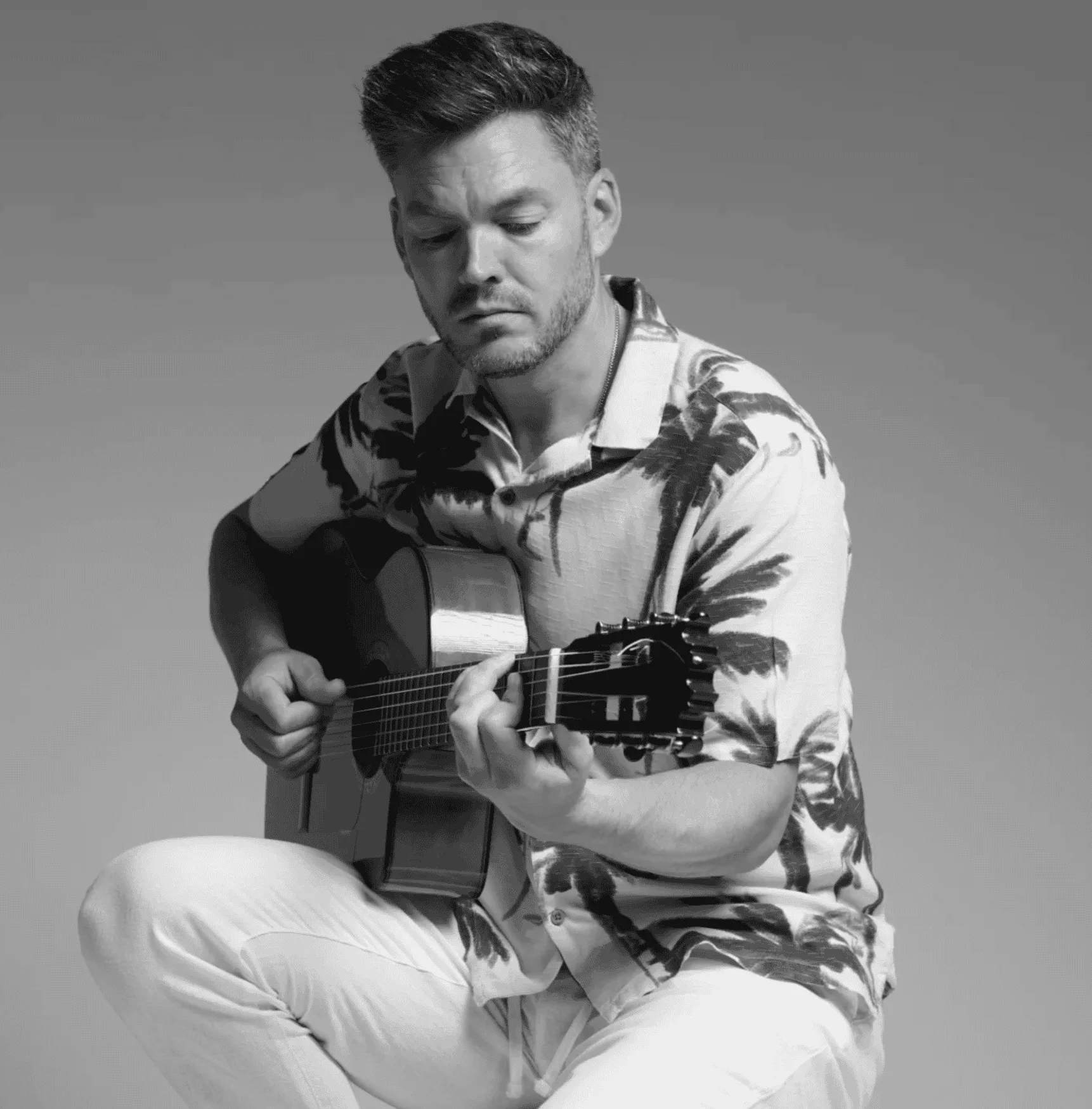 Retrato en blanco y negro de un músico sentado tocando una guitarra española; mira hacia las cuerdas con gesto concentrado. Imagen usada para ilustrar un dúo de rumba fusión en Madrid para la fiesta de tu boda.