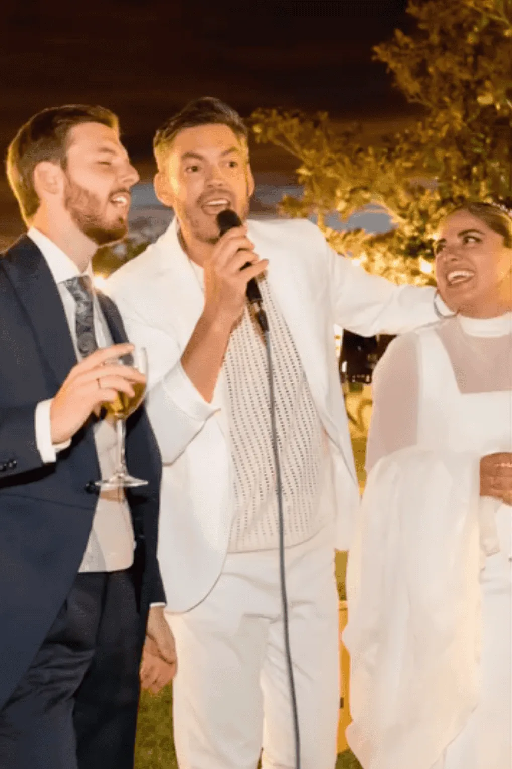 En una celebración de boda al aire libre y de noche, un músico vestido de blanco canta con un micrófono mientras dos invitados a su lado, un hombre con traje azul y copa en la mano y una mujer vestida de blanco, sonríen y le acompañan; escena ideal para un dúo de rumba fusión en Madrid para la fiesta de tu boda.