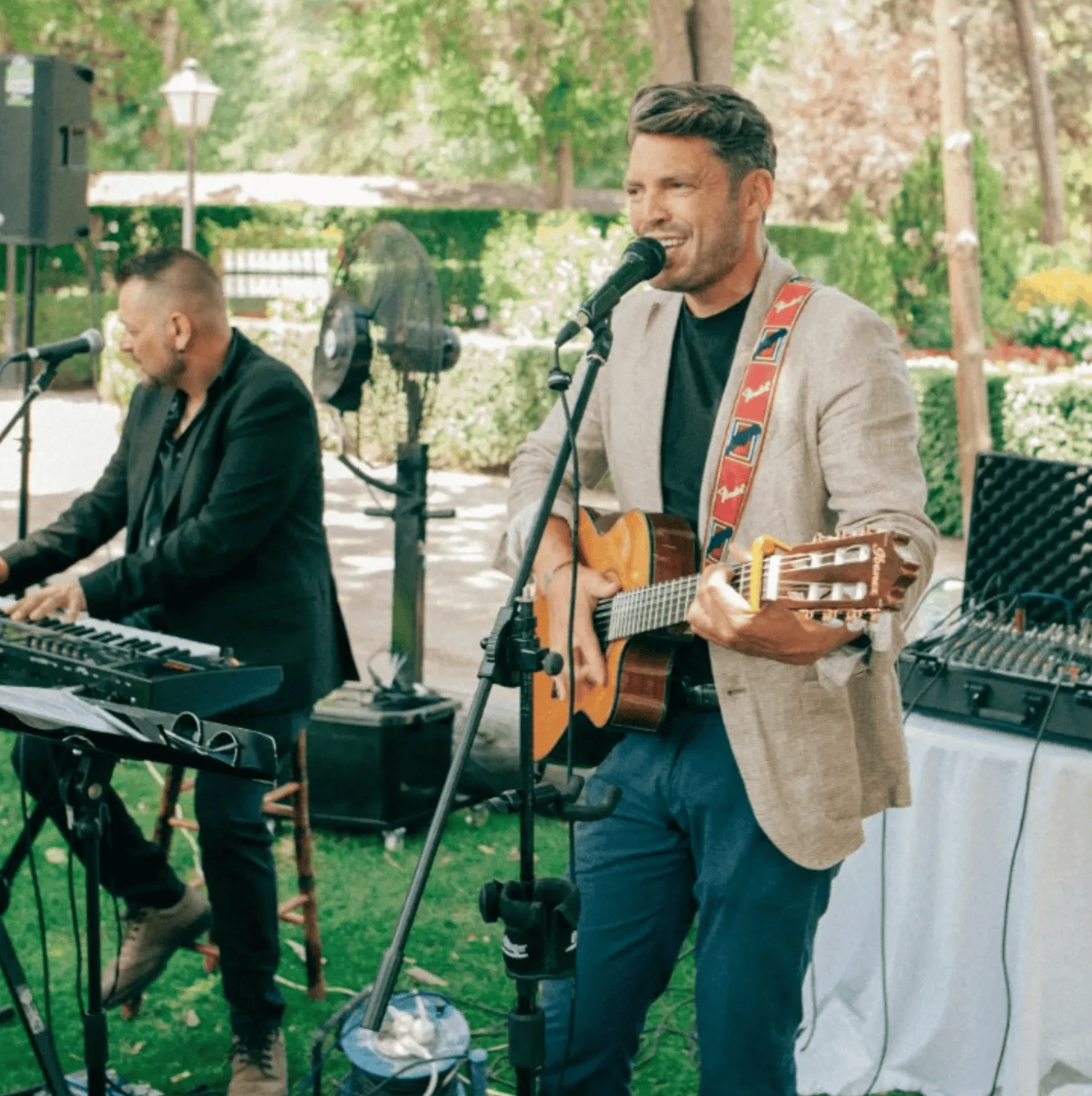 En un jardín, un músico canta sonriendo a un micrófono mientras toca una guitarra española; a su izquierda, otro músico toca un teclado. Hay altavoces y una mesa de sonido, y el ambiente parece el de una celebración al aire libre, ideal como dúo de rumba fusión en Madrid para la fiesta de tu boda.