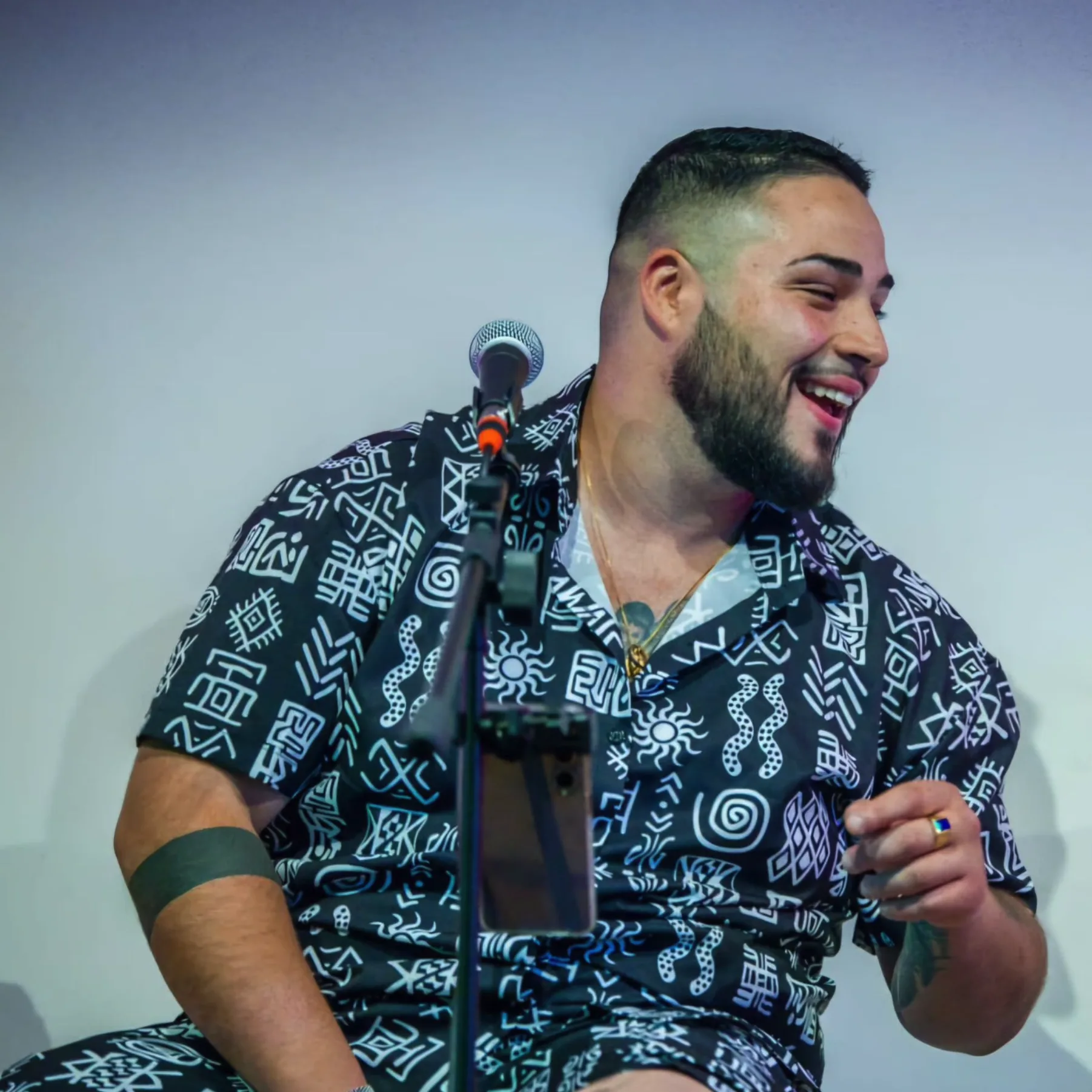 Primer plano de un cantante sonriendo junto a un micrófono en soporte, con camisa negra estampada; imagen pensada para ilustrar un trío de flamenquito en Barcelona para bodas con rollo en terraza.