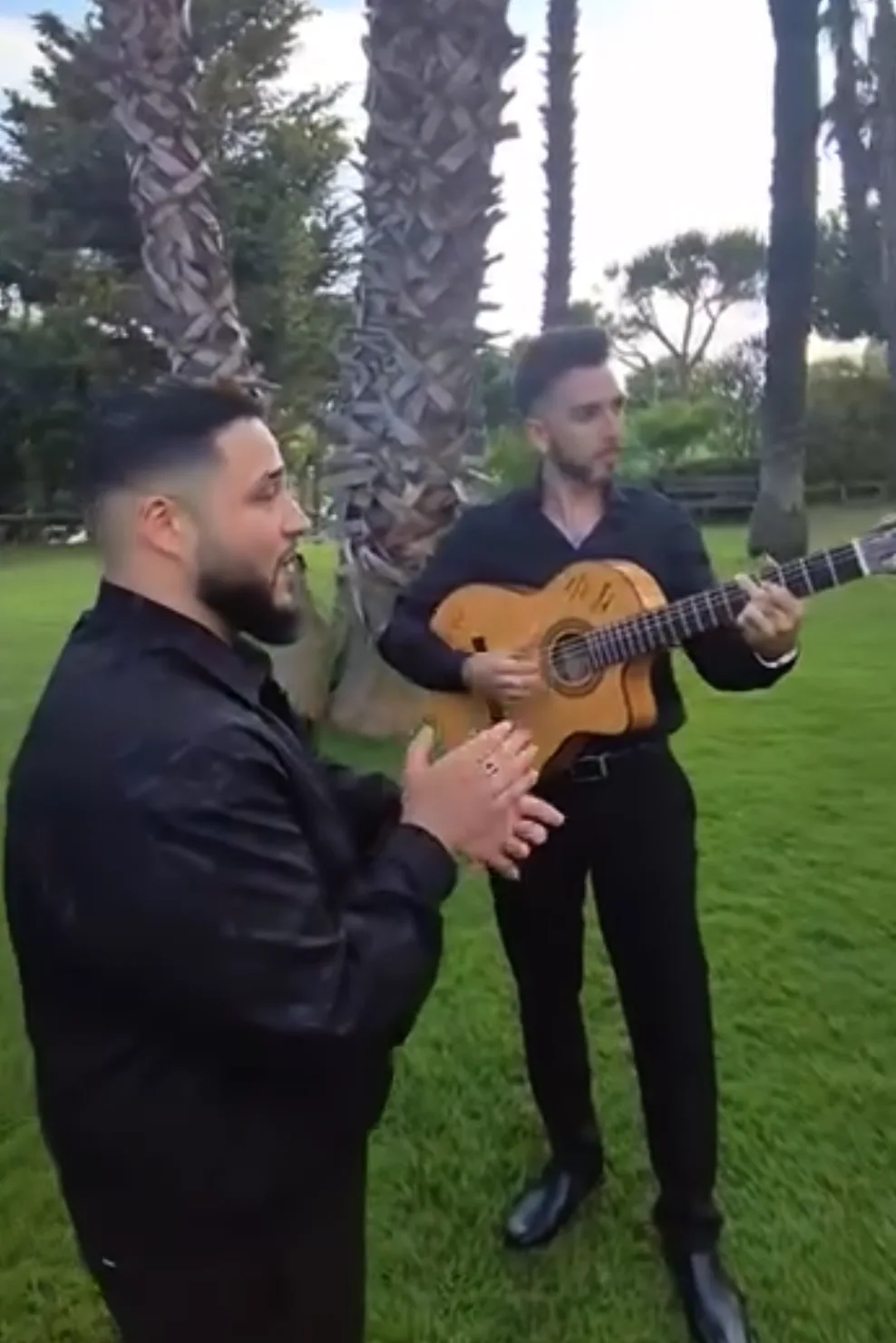 Dos músicos vestidos de negro actúan al aire libre sobre césped: uno toca una guitarra española y el otro acompaña con palmas, con palmeras y jardín de fondo; ambiente de flamenquito pensado para bodas y eventos privados en Barcelona con rollo de terraza.