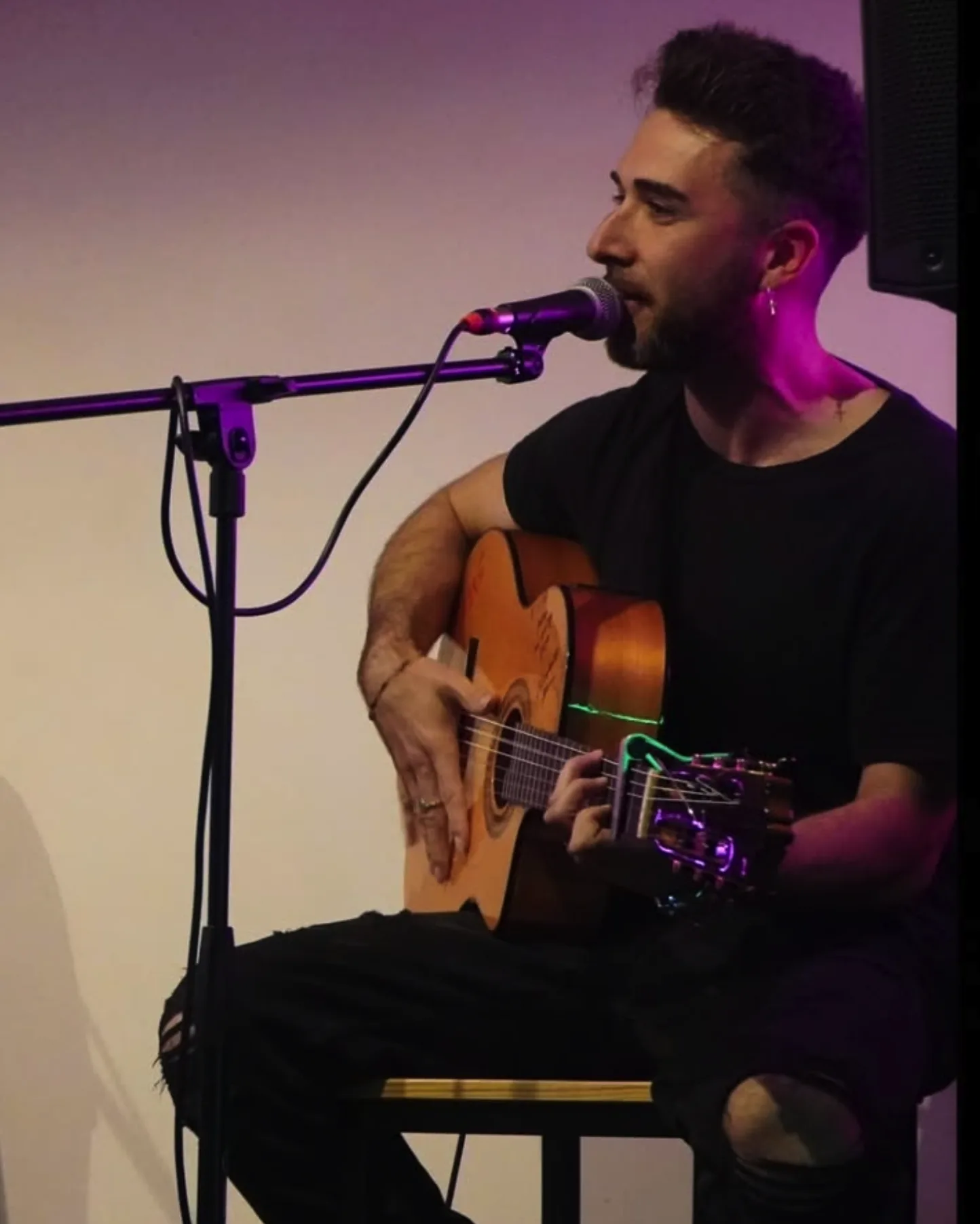 Primer plano de un músico sentado, cantando en un micrófono mientras toca una guitarra española; lleva camiseta negra y un pendiente, con luz violeta sobre el rostro. Escena de estilo flamenquito, pensada para bodas y eventos privados en Barcelona con un ambiente íntimo tipo terraza.
