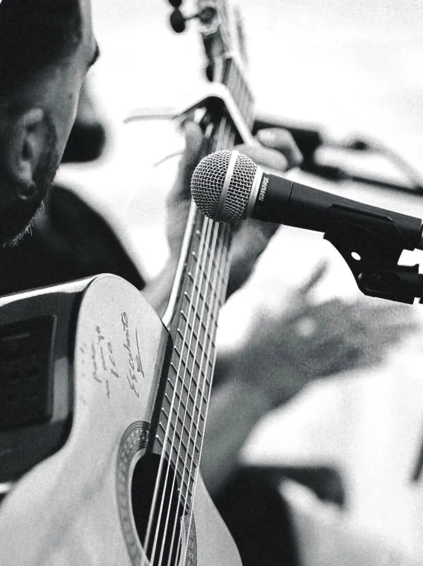 Primer plano en blanco y negro de una guitarra española mientras una mano toca las cuerdas, con un micrófono muy cerca; a la izquierda se ve parcialmente el rostro del músico. Imagen asociada a un trío de flamenquito en Barcelona para bodas con rollo en terraza.