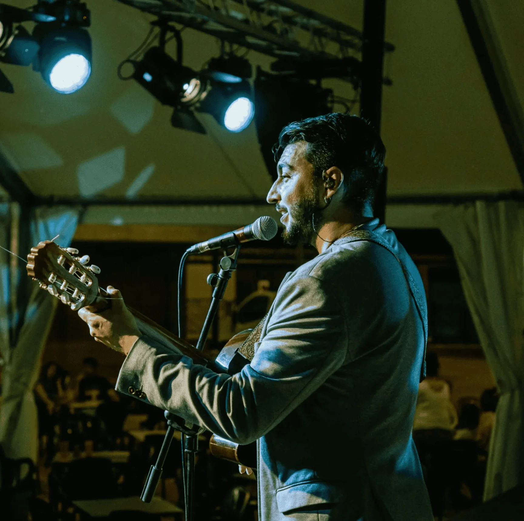 Cantante de perfil con auricular, tocando una guitarra española y cantando en un micrófono bajo focos de escenario; imagen pensada para rumba fusión en Madrid para poner a la gente a tono en tu boda o en celebraciones privadas.
