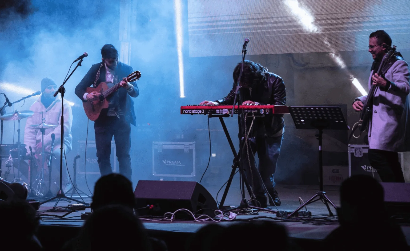 Grupo de música sobre un escenario con humo y luces: un batería a la izquierda, un guitarrista en el centro, un teclista inclinado sobre un teclado rojo y un bajista a la derecha; una escena ideal de rumba fusión en Madrid para poner a la gente a tono en tu boda o celebración.