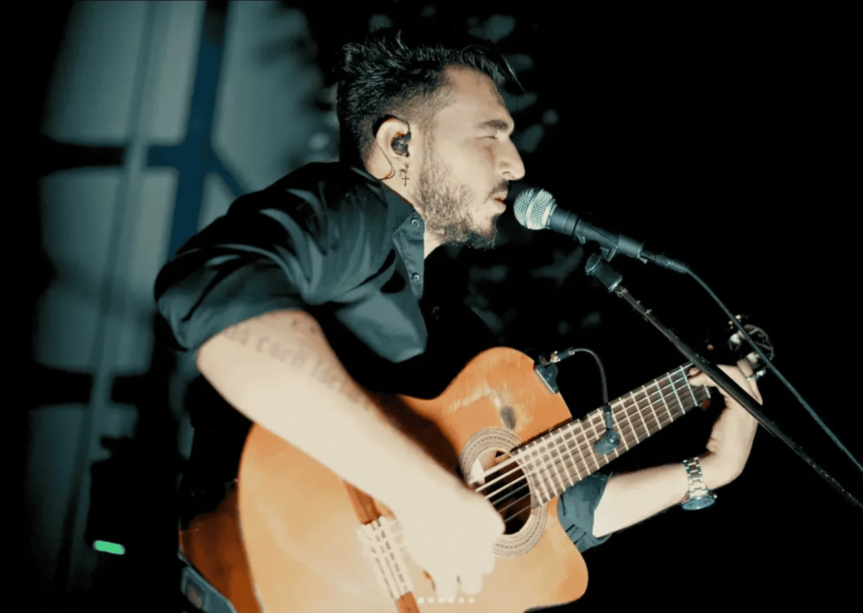 Primer plano lateral de un cantante con auricular in-ear, cantando en un micrófono mientras toca una guitarra española; escena con luz tenue y fondo oscuro, ideal para rumba fusión en Madrid para poner a la gente a tono en tu boda.