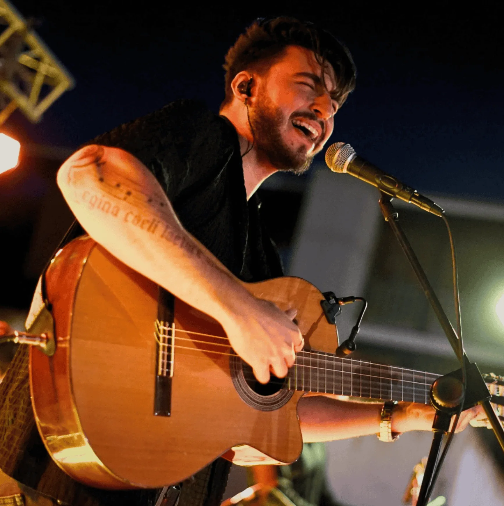 Primer plano de un cantante con barba cantando con los ojos cerrados frente a un micrófono mientras toca una guitarra acústica, iluminado por focos cálidos sobre un escenario; imagen ideal para hablar de rumba fusión en Madrid para poner a la gente a tono en tu boda.