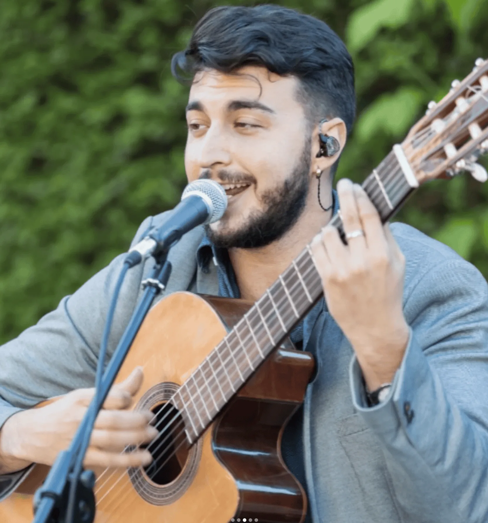 Primer plano de un guitarrista cantando a un micrófono mientras toca una guitarra española; lleva un auricular intrauditivo y aparece con fondo de vegetación desenfocado, una imagen ideal para hablar de rumba fusión en Madrid para poner a la gente a tono en tu boda.