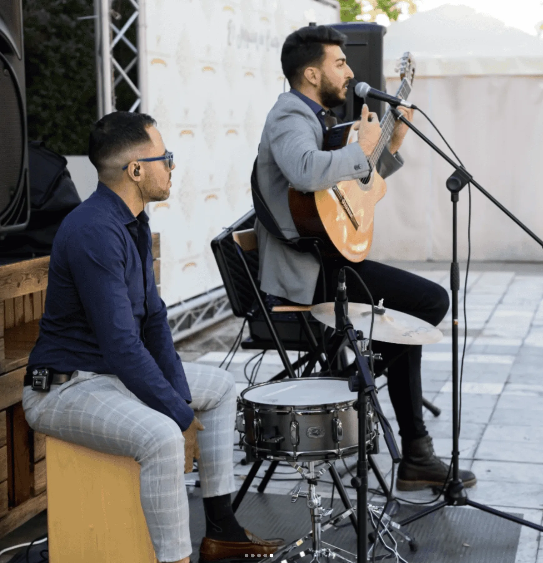 En un pequeño escenario exterior, un músico sentado canta al micrófono mientras toca una guitarra española; a su lado, otro músico con gafas y auriculares se sienta junto a una caja y un plato de batería. La escena encaja con una propuesta de rumba fusión en Madrid para poner a la gente a tono en tu boda.