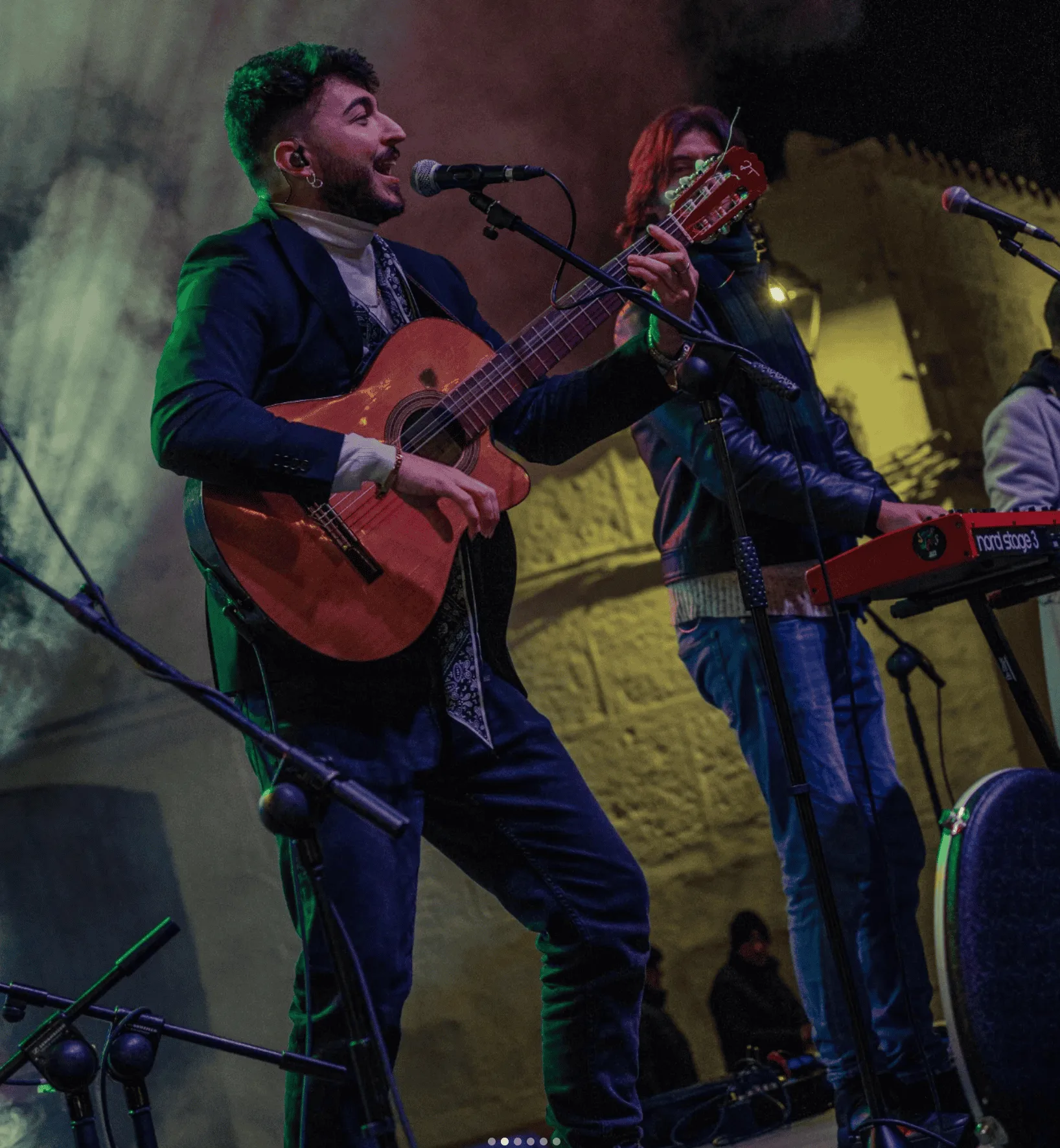 En un escenario con luces de colores, un cantante con chaqueta canta al micrófono mientras toca una guitarra española; detrás, otro músico toca un teclado. Imagen de rumba fusión en Madrid pensada para poner a la gente a tono en tu boda.