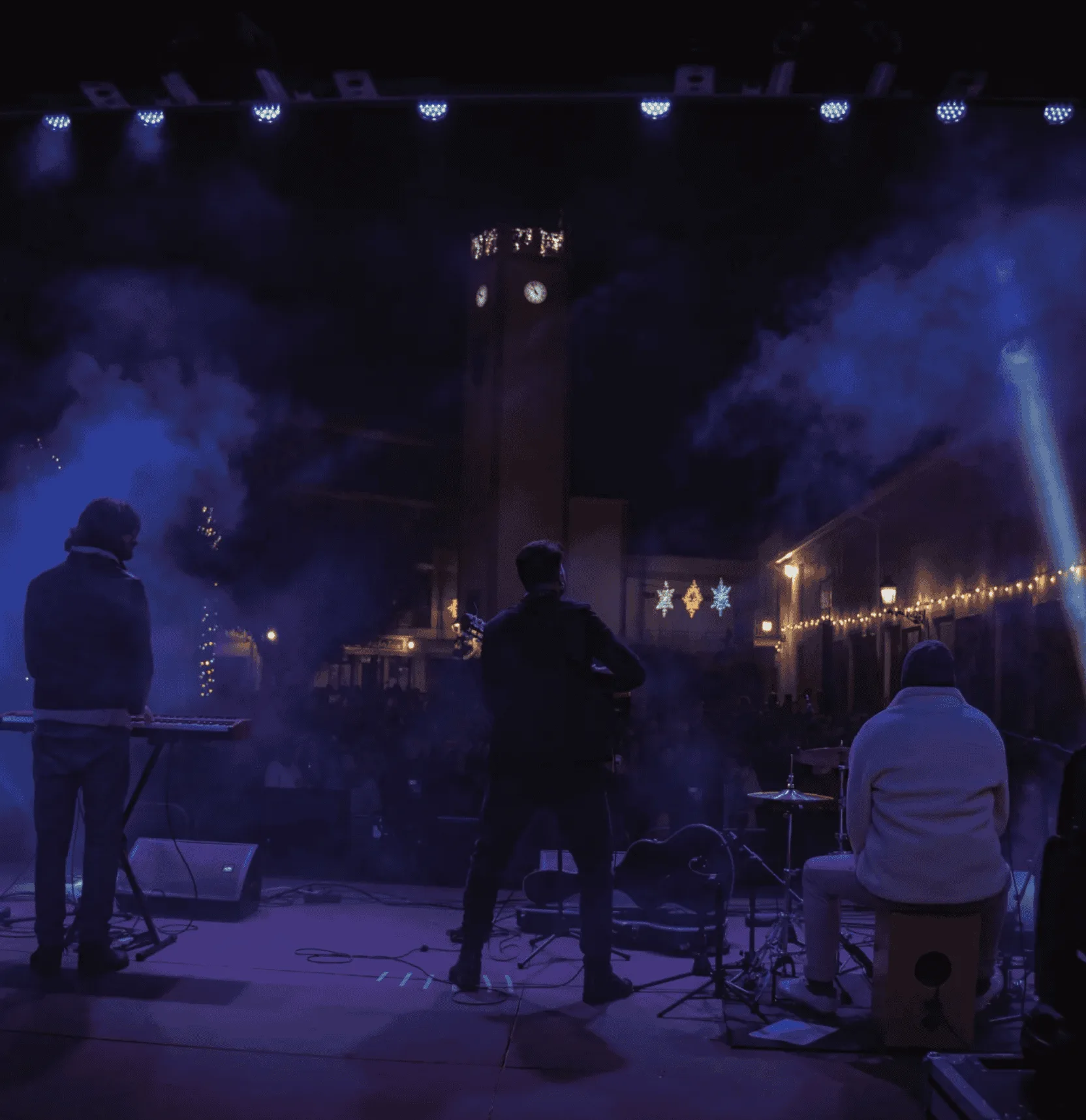 Vista desde detrás del escenario: tres músicos de espaldas con instrumentos y una batería en primer plano, bajo focos azules y humo, mientras el público se intuye al fondo en un recinto exterior iluminado; ambiente ideal para una rumba fusión en Madrid para poner a la gente a tono en tu boda.
