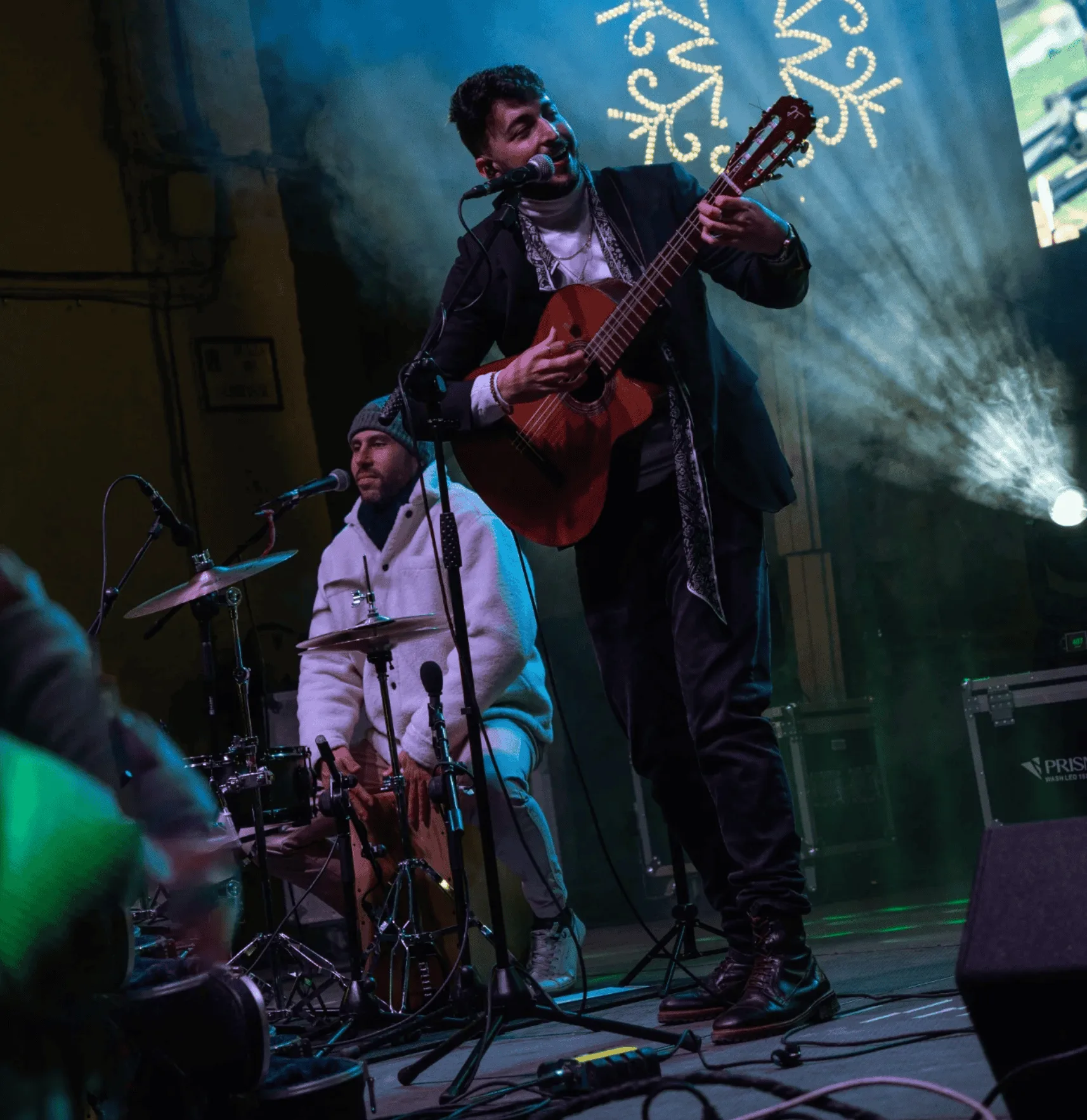 En un escenario con luz azul y humo, un músico canta sonriendo a un micrófono mientras toca una guitarra española; a su lado, un batería sentado acompaña el ritmo. Imagen de rumba fusión en Madrid para poner a la gente a tono en tu boda o en un evento privado.