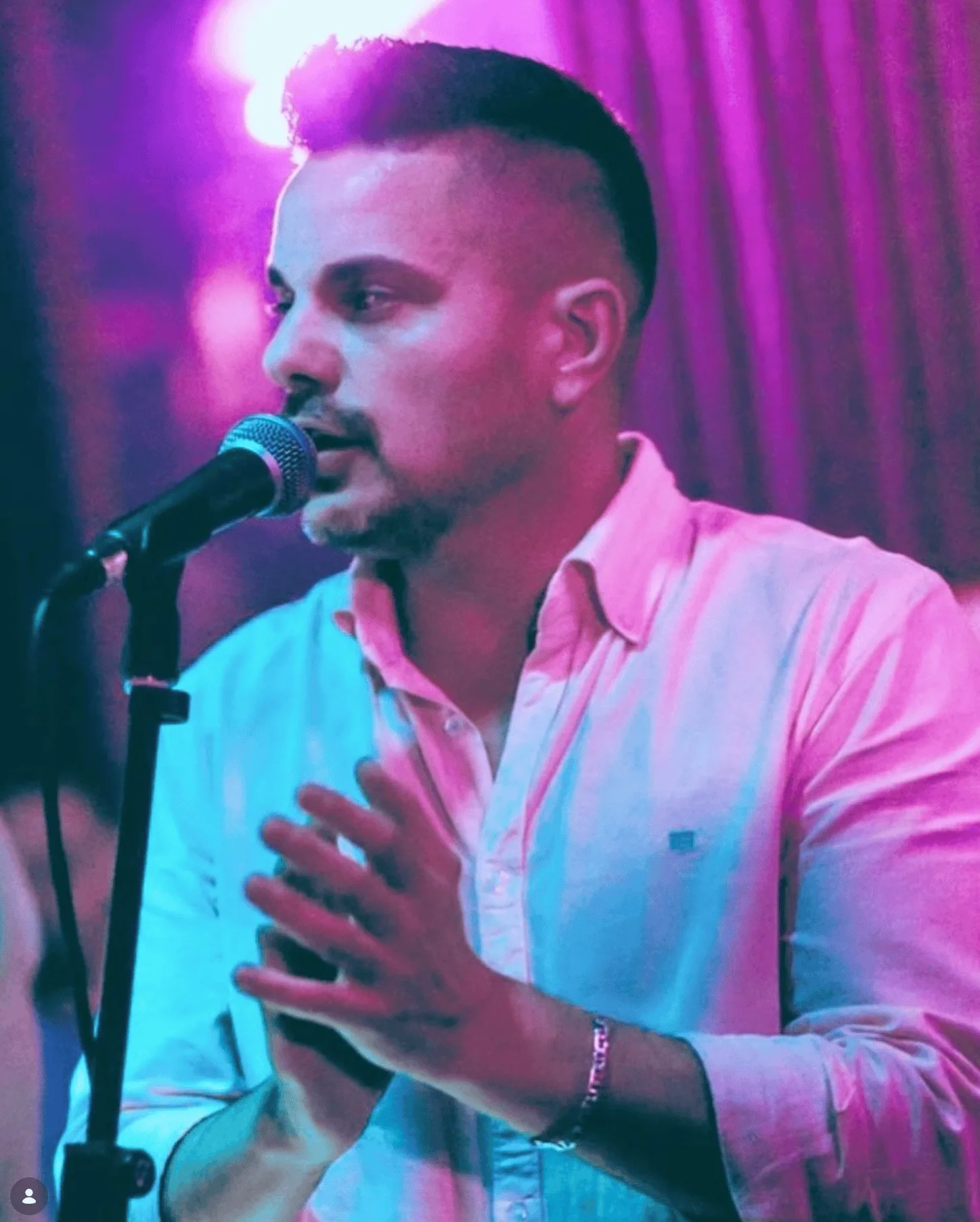 Primer plano de un cantante con camisa clara, cantando en un micrófono sobre un soporte mientras gesticula con una mano; el fondo está desenfocado con iluminación rosa y morada, una escena típica para rumba fusión y flamenquito en bodas y Fallas en Valencia.