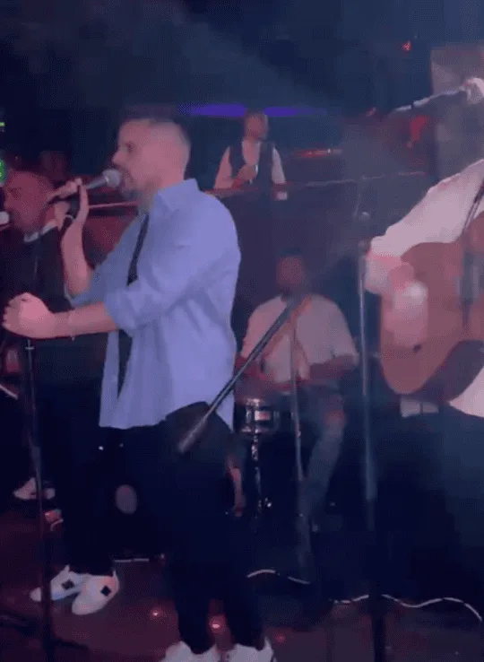 En un escenario interior con luz tenue, un cantante sostiene un micrófono y canta mientras, a su derecha, otro músico toca una guitarra; al fondo se aprecia un percusionista sentado y parte del público. Imagen de rumba fusión y flamenquito pensada para bodas y Fallas en Valencia.