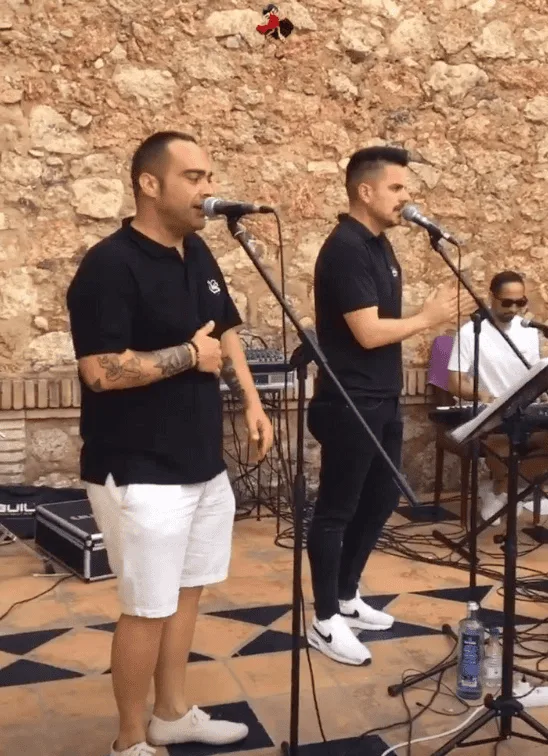 Dos cantantes con micrófonos en pie actúan frente a una pared de piedra; a la derecha, un teclista con gafas toca sentado. El grupo viste ropa oscura y están montados sobre un suelo de baldosas, con cables y equipo de sonido alrededor, en un ambiente de celebración ideal para rumba fusión y flamenquito para bodas y Fallas en Valencia.