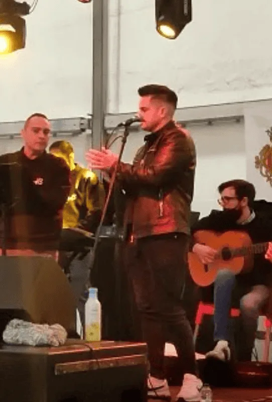 Cantante con chaqueta de cuero aplaudiendo frente a un micrófono en un escenario interior, mientras un guitarrista sentado toca a su lado; iluminación cálida y ambiente de actuación, ideal para rumba fusión y flamenquito para bodas y Fallas en Valencia.
