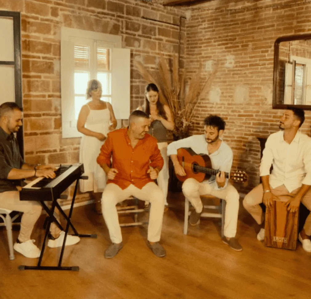 Grupo de seis personas en una sala interior con paredes de ladrillo: un músico toca un teclado a la izquierda, otro toca una guitarra española sentado en el centro y un tercero toca un cajón a la derecha; en medio, un hombre con camisa roja marca el ritmo sentado mientras dos mujeres de pie observan detrás. Imagen que ilustra un trío de pop español y rumba para fiestas mayores en Barcelona, con ambiente cercano y festivo.