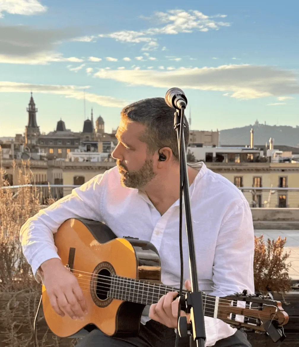 Primer plano de un guitarrista con camisa blanca sentado junto a un micrófono, tocando una guitarra española en una azotea; al fondo se ven edificios de Barcelona bajo un cielo con nubes. Imagen para ilustrar un trío de pop español y rumba para fiestas mayores en Barcelona.