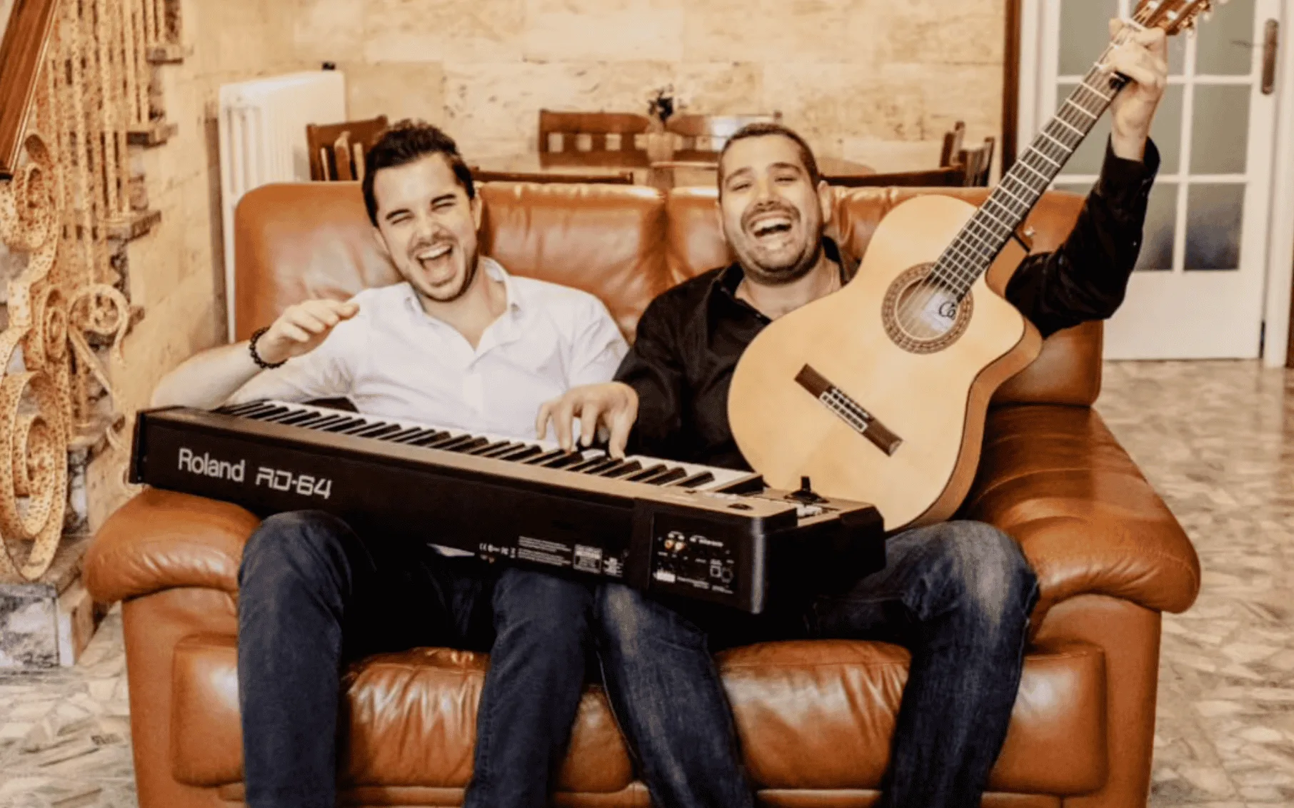 Dos músicos sentados en un sofá de cuero en un interior cálido; uno sostiene una guitarra española y sonríe con el brazo levantado, y el otro tiene un teclado Roland apoyado sobre las piernas. Imagen promocional de un trío de pop español y rumba para fiestas mayores en Barcelona.