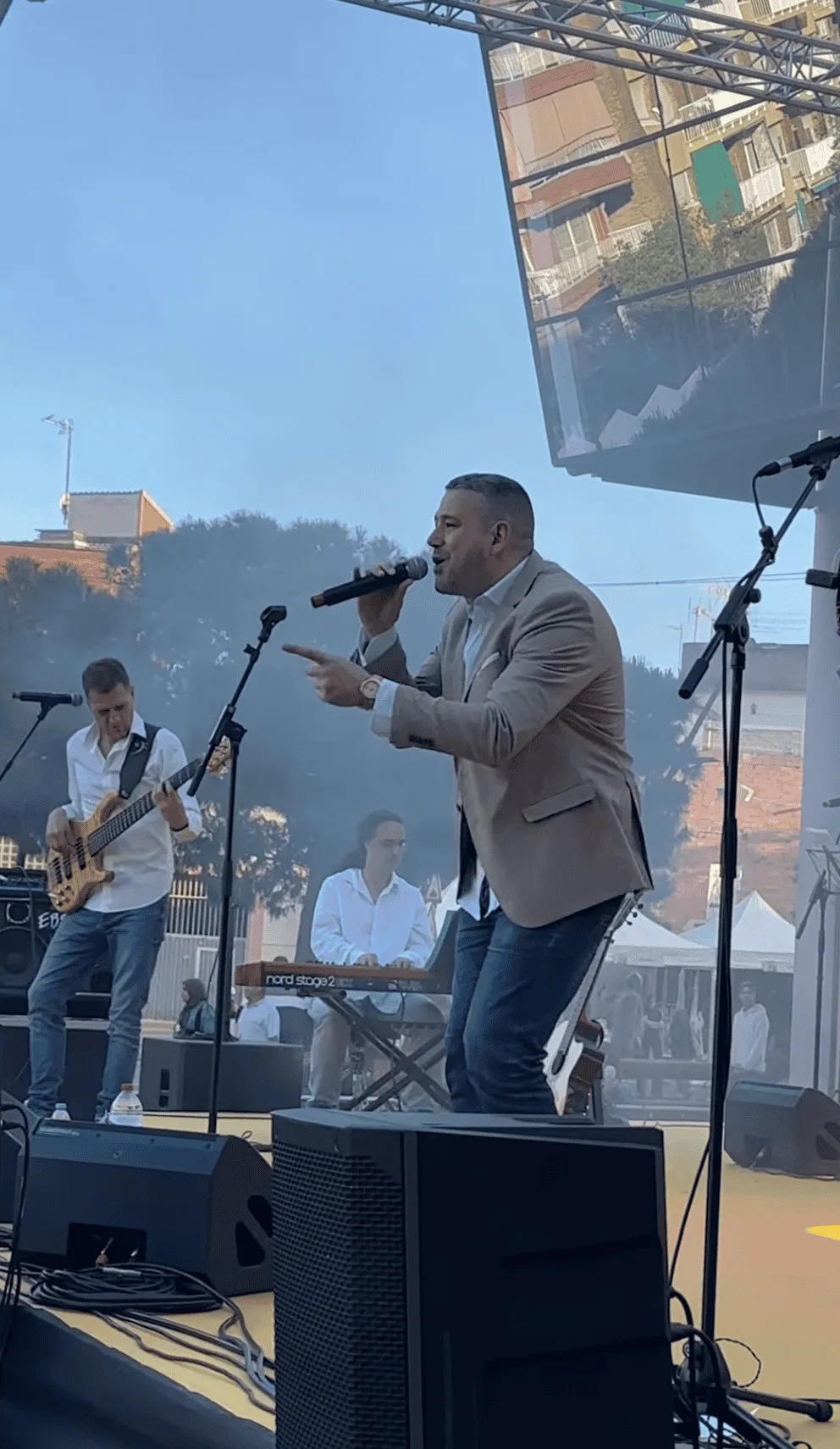 En un escenario al aire libre, un cantante con micrófono en la mano y chaqueta beige actúa mientras a su izquierda un bajista toca y, al fondo, un teclista acompaña. Se ven varias peanas de micro, altavoces en primer plano y una gran pantalla con edificios alrededor, en un ambiente de fiesta de calle en Barcelona; imagen representativa de un trío de pop español y rumba para fiestas mayores en Barcelona.