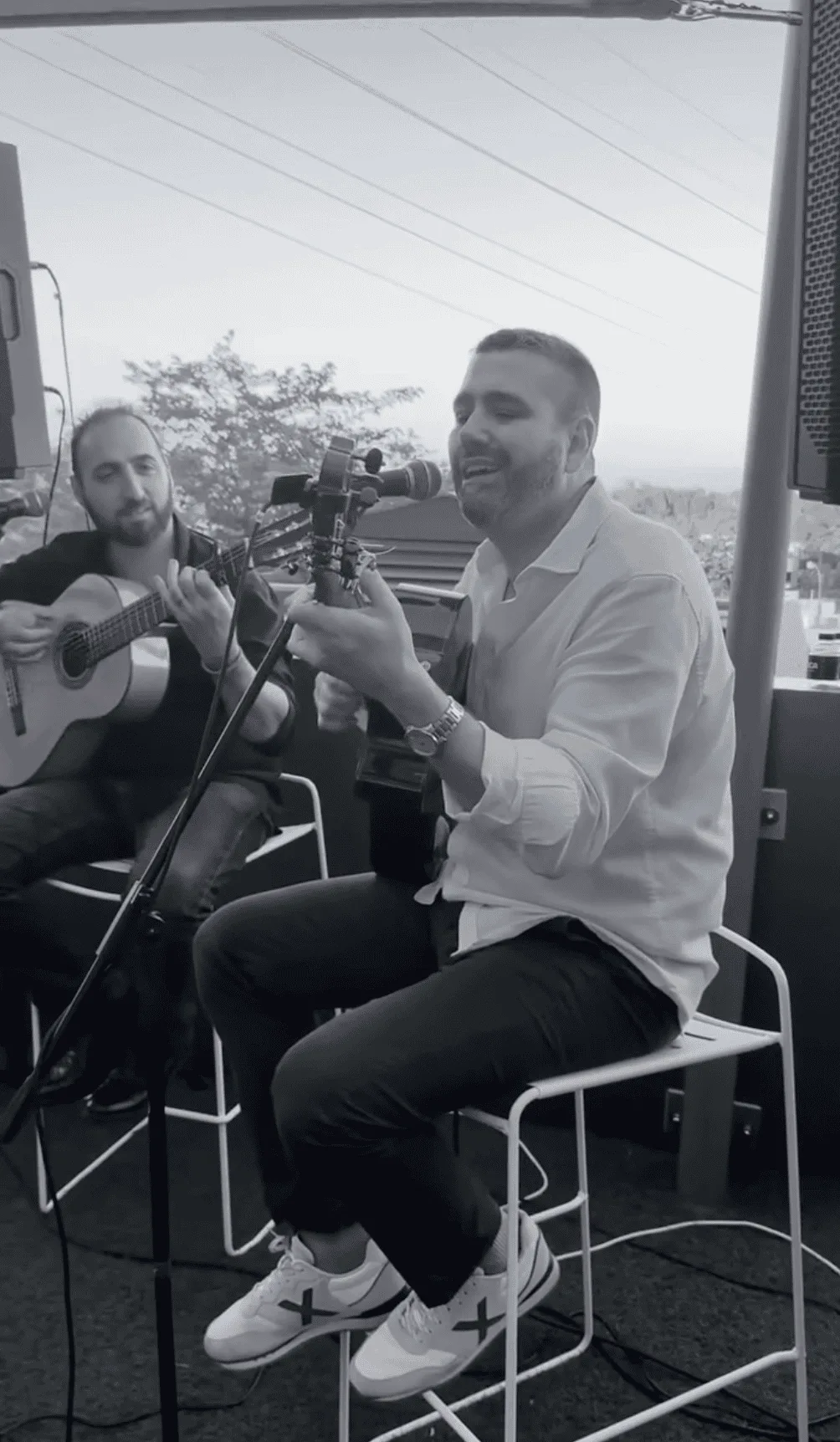 Dos músicos sentados en un escenario al aire libre: uno canta frente a un micrófono mientras toca la guitarra y el otro acompaña con guitarra española al fondo; imagen en blanco y negro con equipo de sonido, ideal para un trío de pop español y rumba para fiestas mayores en Barcelona.