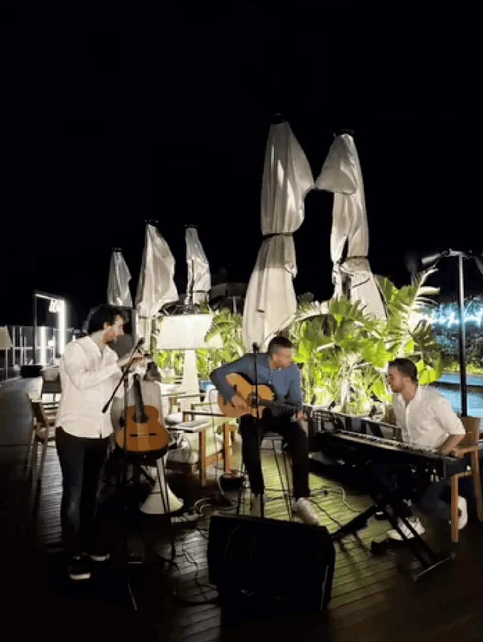 Terraza al aire libre de noche con un trío musical: un cantante de pie junto a un micrófono, un guitarrista sentado con guitarra acústica y un pianista en un teclado; alrededor hay cables, altavoces y sombrillas cerradas, en un ambiente de evento. Imagen de un trío de pop español y rumba para fiestas mayores en Barcelona.