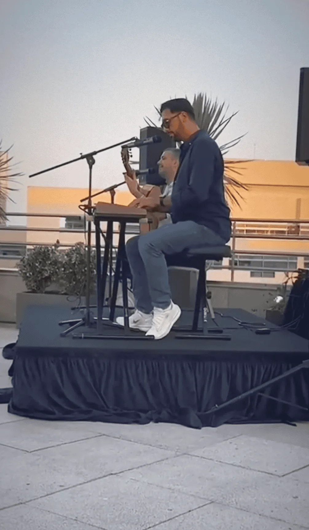 Dos músicos sentados en un pequeño escenario al aire libre: en primer plano un hombre con gafas canta a un micrófono mientras toca una caja tipo percusión sobre las piernas, y detrás otro músico aparece con una guitarra; a ambos lados hay altavoces y al fondo se ve un edificio y una palmera, ambiente propio de un trío de pop español y rumba para fiestas mayores en Barcelona.