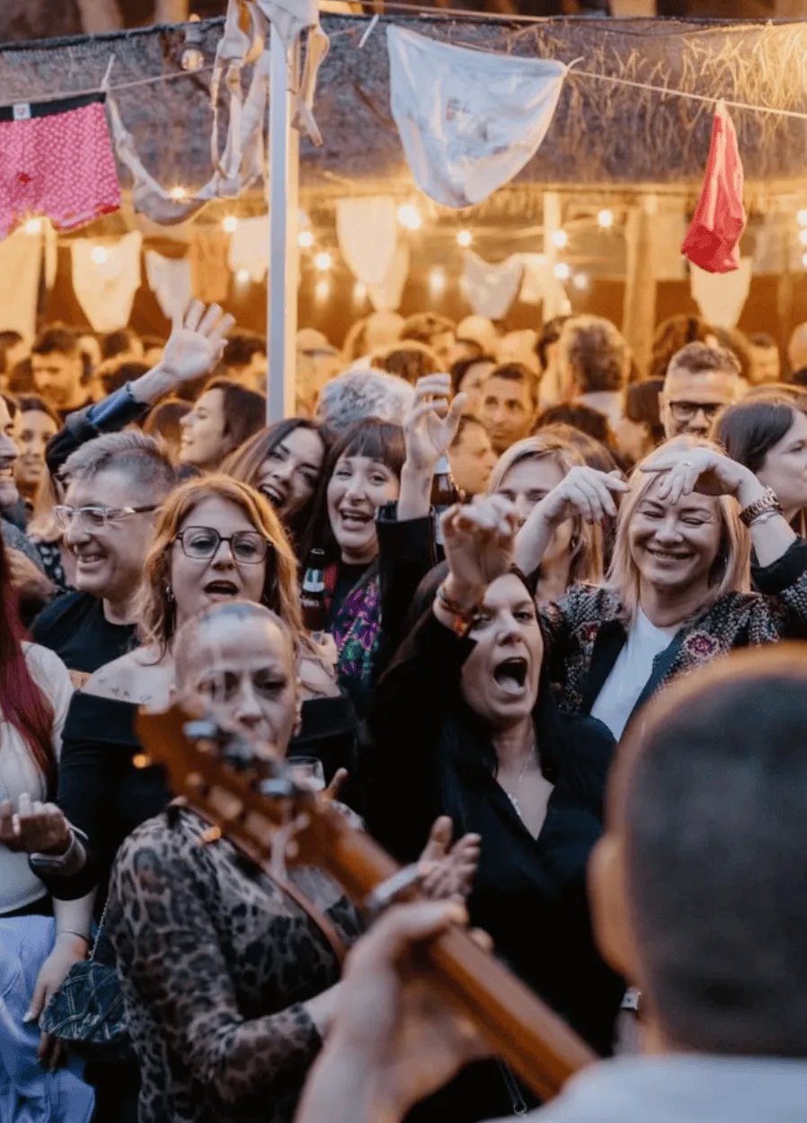 Grupo de personas bailando y cantando en una fiesta nocturna al aire libre, con guirnaldas de luces y pañuelos colgados; en primer plano se ve desenfocado el mástil de una guitarra, creando ambiente de trío de pop español y rumba para fiestas mayores en Barcelona.