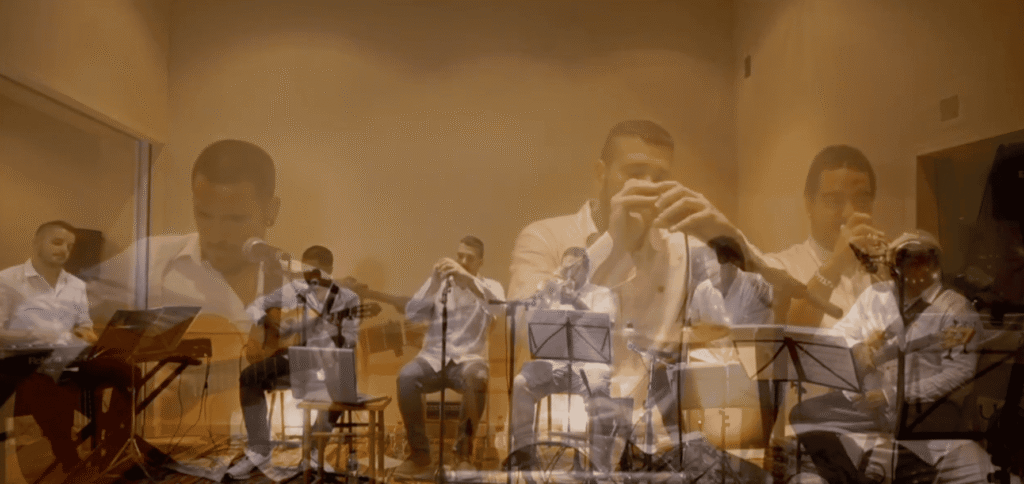 Grupo de músicos num salão interior com luz quente, sentados e a tocar em conjunto: um teclista à esquerda, um cantor ao microfone e vários instrumentistas com sopros, com partituras em suportes à frente; a imagem tem um efeito de sobreposição que destaca um dos músicos em primeiro plano. Ideal para ilustrar um trio de pop espanhol e rumba para festas maiores em Barcelona.