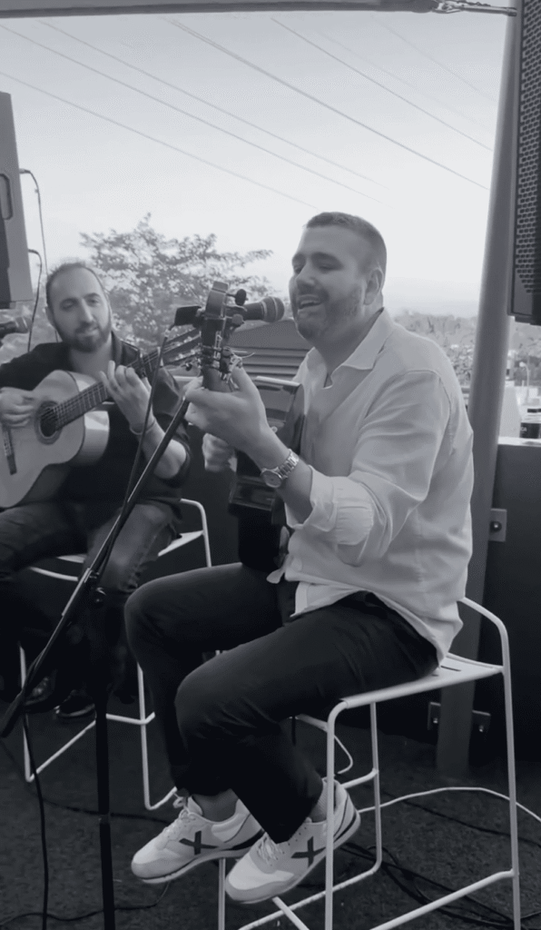 Dois músicos sentados num terraço, em foto a preto e branco, a tocar guitarra espanhola e a cantar num microfone, com colunas ao lado e vista de árvores e cidade ao fundo, num ambiente de celebração ideal para quem procura um trio de pop espanhol e rumba para festas maiores em Barcelona.